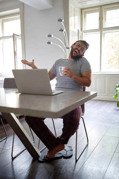 Junger gutaussehender Mann mit Bart und Brille, lächelnd und telefonierend, während er am Tisch sitzt und auf dem Laptop — Stockfoto