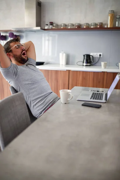 Volwassen jongeman met baard en tatoeage, het dragen van een bril, gazons, stretching in de voorkant van laptop op tafel. — Stockfoto