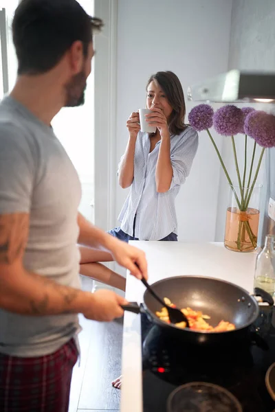 Joven pareja en cocina hablando, sonriendo, mientras que cocinar y beber café . —  Fotos de Stock