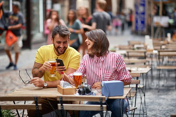 Homem e menina sentados em um café bebendo cerveja e olhando para fotos em um telefone celular . — Fotografia de Stock