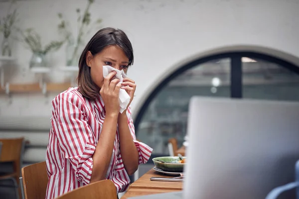 Sick woman at caffe working on laptop. Blowing nose on tissue. — Stock Photo, Image