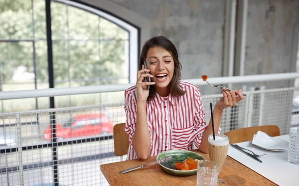 Beautiful young brunette talking on a cell phone in restaurant while eating. — Stock Photo, Image