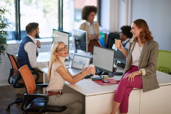 two female colleagues talking, drinking coffee and smiling at work, with group of colleagues in the background. casual, open space, modern, office, business concept