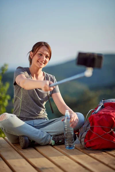 female enjoying in nature, sitting with legs crossed at viewpoint high in mountain looking at cell phone, taking selfie. nature, relaxing, leisure, positive, energy concept