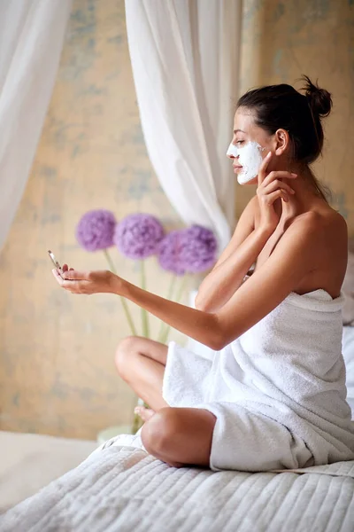 年轻美丽的女性坐在床上 看着小镜子 从她的脸上摘下一个美丽的面具 — 图库照片