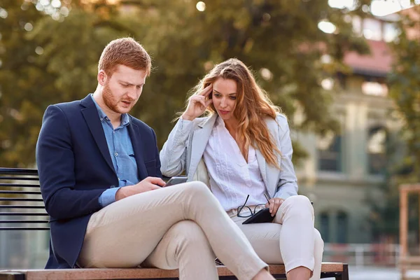 若い白人ビジネスマンが公園のベンチで女性同僚に携帯電話でコンテンツを見せる コロナウイルスのロックダウンの概念の後で真剣に心配しているビジネスマン — ストック写真