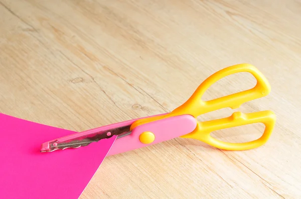 Uma tesoura colorida que corta um padrão ziguezague com um papel rosa — Fotografia de Stock