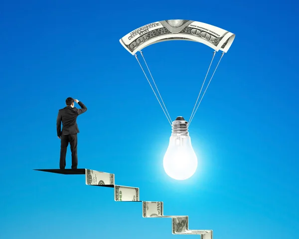 Человек на денежной лестнице смотрит лампочку с денежным парашютом — стоковое фото