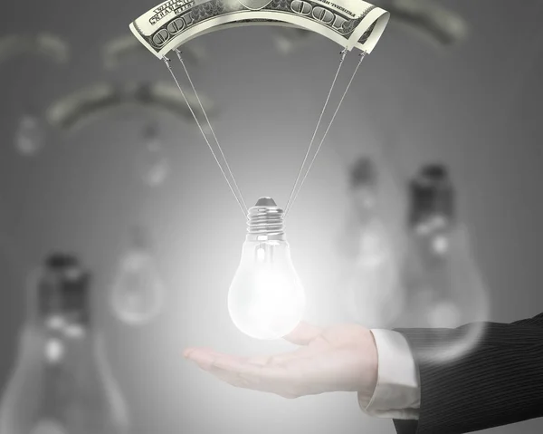 Human hand holding bright light bulb with money parachute. — Stock Photo, Image