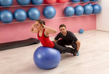 Koçunun yardım ettiği genç bir kadın spor salonunda spor topuyla egzersiz yapıyor..