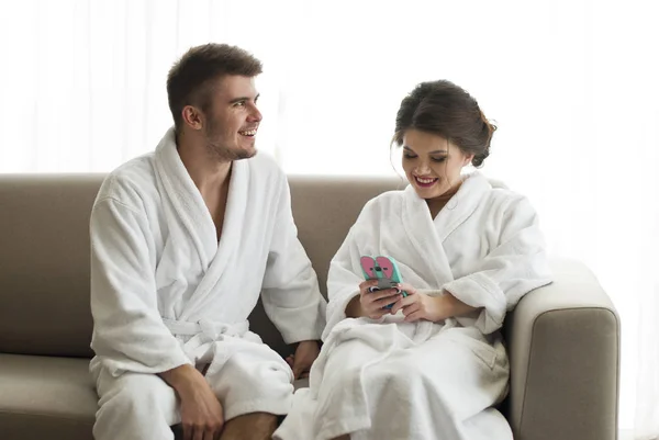 魅力的な若いカップルはバスローブでソファの上のスマート フォンを見ています。 — ストック写真