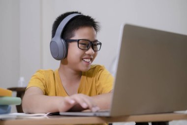 Neşeli çocuk internetten bilgisayar kullanarak okul ödevlerini öğrendiği için heyecanlı. Asyalı çocuk evde e-öğrenme ile kendi kendine çalışmanın keyfini çıkarıyor. Çevrimiçi eğitim ve kendi kendine öğrenim ve evde eğitim kavramı.