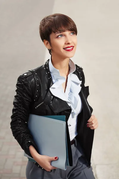 Happy young fashion business woman in leather jacket with a folders walking in city street