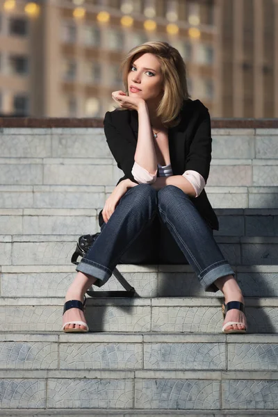 Young blonde fashion business woman sitting on steps Stylish female model in black blazer and dark blue jeans
