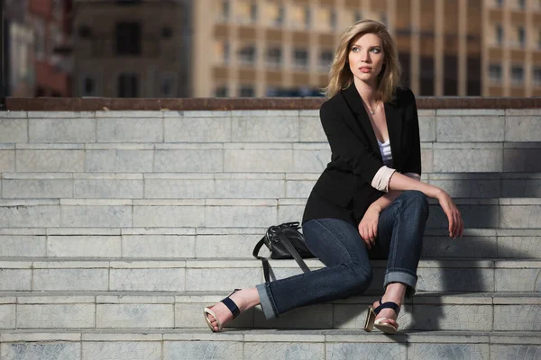 Young blonde fashion business woman sitting on steps Stylish female model in black blazer and dark blue jeans