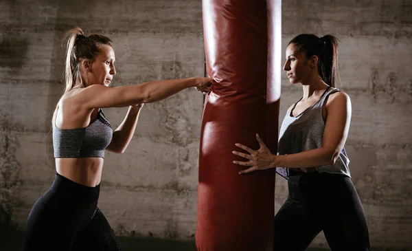 Young muscular woman punching a boxing bag on cross fit training with a female personal trainer at the gym.