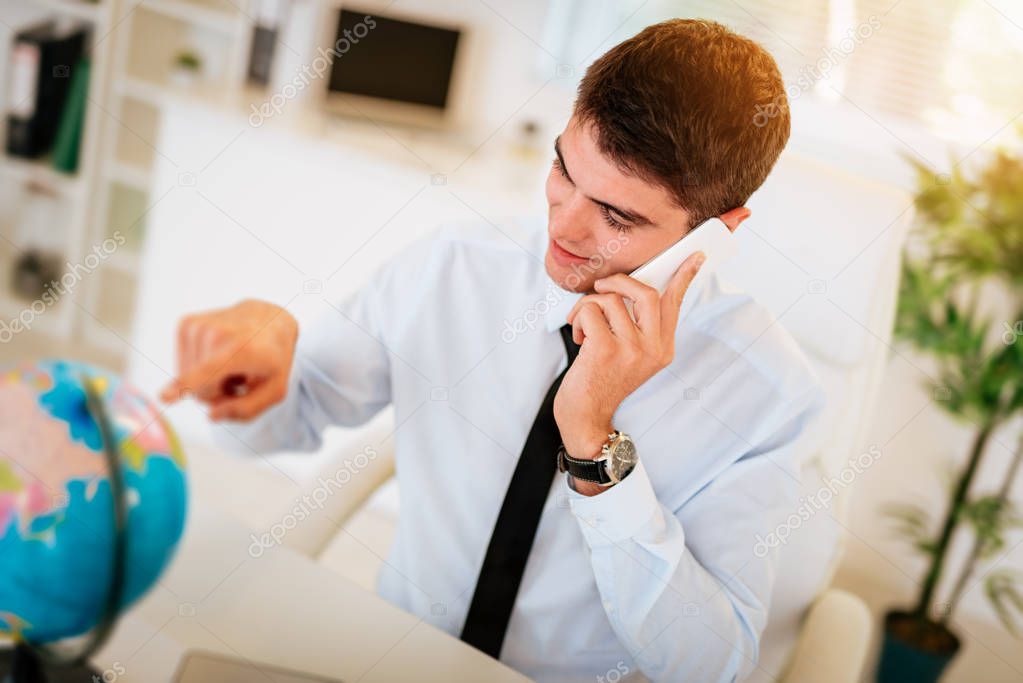 Young businessman using smartphone and looking at globe at discovering available markets of global business. He pointing finger on the globe on the table in front of him.