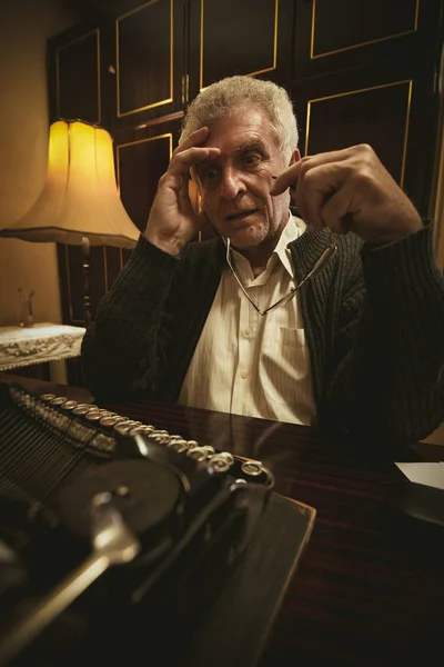 古いタイプライターおよび思考の側の机に座って 彼の手で眼鏡をかけて物思いにふけるレトロなシニア男性作家 — ストック写真