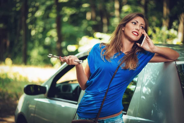 美丽的年轻女子 车在森林中的道路上崩溃 她站在车旁 手持扳手 在手机上说话 — 图库照片