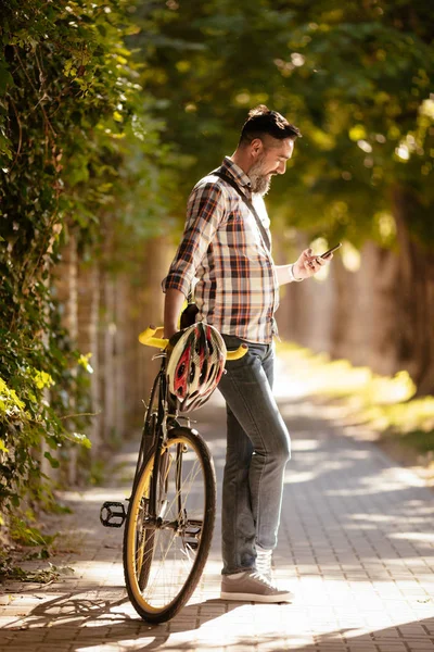 ハンサムな実業家は 自転車で仕事に行きます 彼はバイクの横に立って スマート フォンを使用して — ストック写真