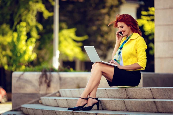 成功的女商人坐在办公区的楼梯上 在笔记本电脑上工作 谈论智能手机 — 图库照片