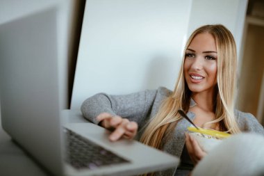 Kahvaltı ve dizüstü bilgisayar kullanarak yatağın üstüne oturan çekici gülümseyen kız. O net sörf ve sosyal medya kontrol. 