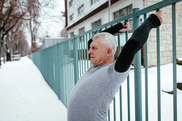 Active senior man stretching and doing exercises using the fence during the winter training outside in.