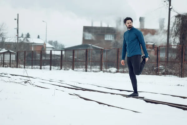 Active young jogging man relaxing legs muscles after training in ghetto during winter training outside.