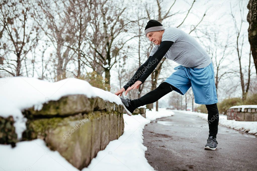 Active senior man stretching before jogging in public park during winter training outside.