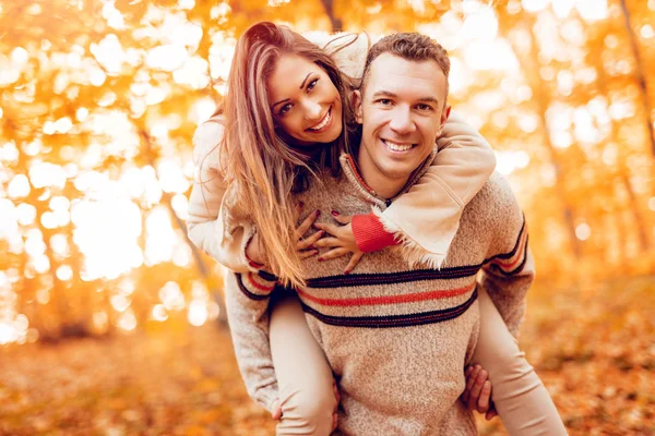 一个年轻的笑脸情侣的肖像在阳光明媚的森林里享受着背驮在秋天的颜色和看相机 — 图库照片