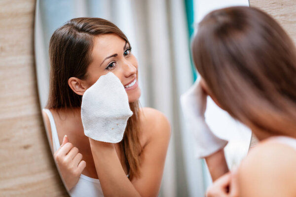 smiling young woman removing make up in front of mirror in bathroom