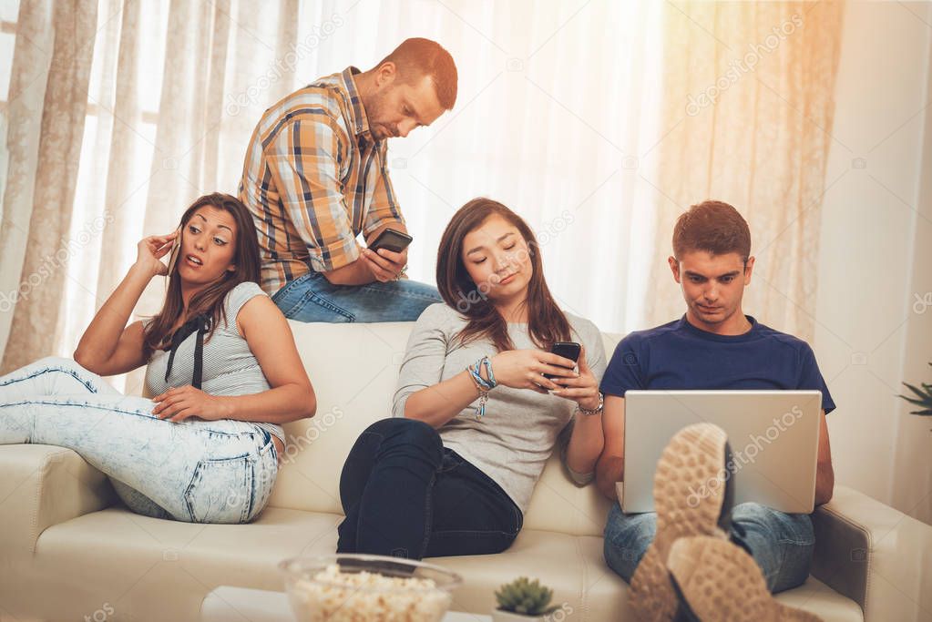 Four bored friends hanging out in an apartment and using smartphones and laptop