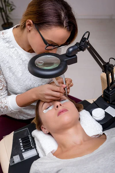 Beautician applying extended eyelashes to young woman in beauty-salon