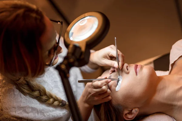 Beautician applying extended eyelashes to young woman in beauty-salon