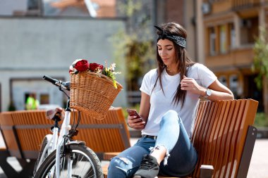 Genç kadın manifatura smartphone ve çiçek sepeti ile Bisiklet yanında güneşli bir günde bir bankta oturan gülümseyen.