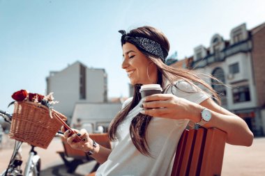 Smartphone, alma kahve molası ve çiçek sepeti ile Bisiklet yanında güneşli bir günde bir bankta oturur gülümseyen genç kadın.