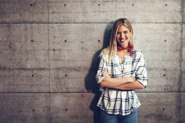 Çapraz elle beton bir duvarı önünde duran ve kameraya bakarak genç güzel başarılı gülümseyen kadın girişimci.