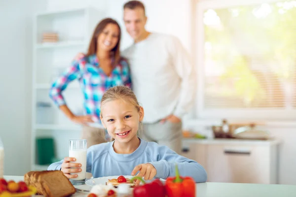 Hermosa Niña Desayunando Cocina Doméstica Sus Padres Felices Pie Detrás —  Fotos de Stock