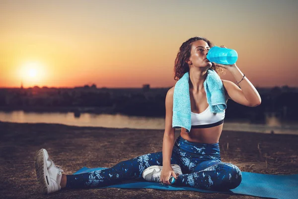 Young fitness woman resting after hard training near river on sunset background