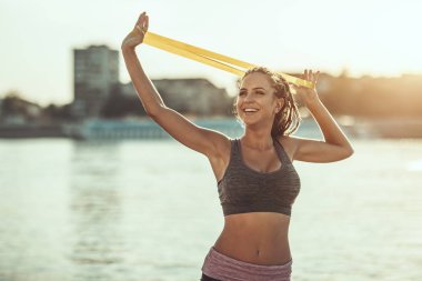 Kaslı genç kadın nehir kıyısında lastik bant ile güçlü bir vücut çalışması yapıyor..