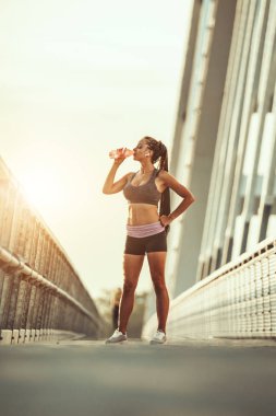 Genç bayan koşucu su içiyor, koşmaya hazırlanıyor ve bir nehir köprüsünde sabah antrenmanı yapıyor..