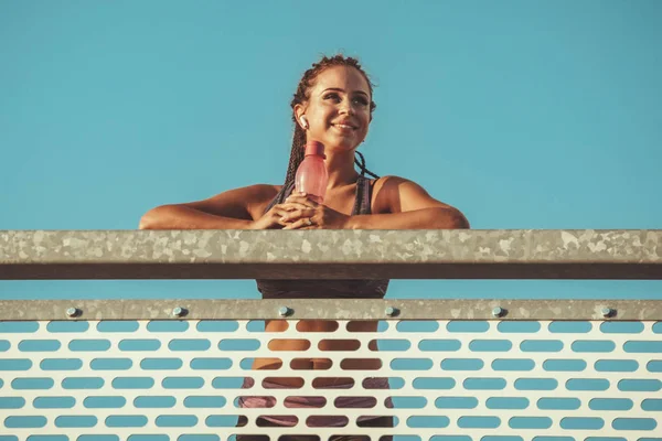 Young fitness woman is resting after hard training on the river bridge, listening music  and looking away.