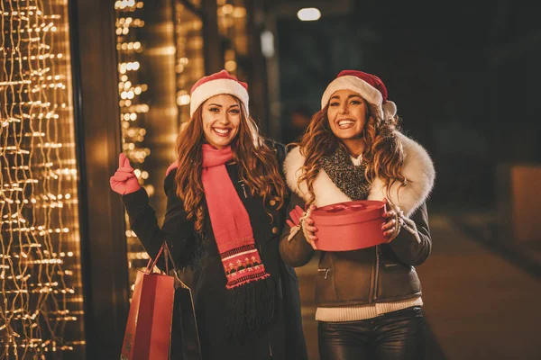 2人の女性の友人は冬の夜を楽しみ 買い物袋と赤い箱を手に笑っています — ストック写真