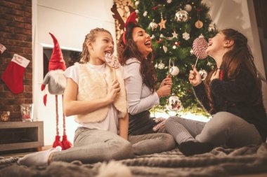 Genç anne ve küçük kızları Noel ağacının yanında oturuyorlar, gülüyorlar ve mutlu görünmekten zevk alıyorlar..