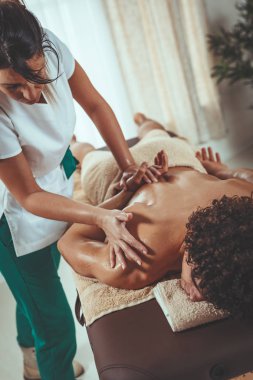 Yakışıklı adam masaj masasında uzanıyor ve masaj merkezinde genç bayan terapist tarafından sağlıklı bir omuz masajı yaptırıyor..