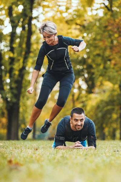 Krásná veselá žena a její partner jsou cvičení v parku, oblečeni v černém obleku s Ems elektronický simulátor stimulovat své svaly. - Stock obrázek