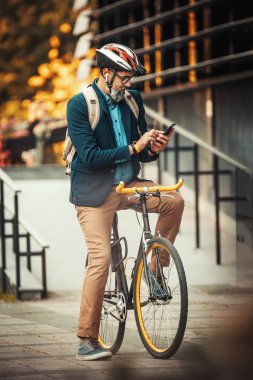 Sıradan yakışıklı orta yaşlı işadamı ofise bisikletle gidiyor. Bisikletin üstünde duruyor ve akıllı telefon kullanıyor..