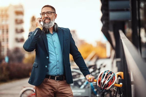A handsome middle-aged man goes to work by bicycle and talking on the smartphone in front of office district.