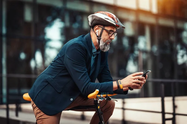 Casual handsome middle-aged businessman is going to work by bicycle. He is standing on the bike and using smart phone in front of office district.