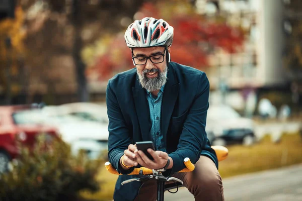 カジュアルでハンサムな中年のビジネスマンは自転車で仕事をしようとしている 彼は自転車に乗っていて スマートフォンを使っている — ストック写真